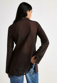 Camicia marrone trasparente a maniche lunghe con design sagomato, caratterizzata da un retro leggermente allungato e una texture liscia. Jeans blu visibili sotto.