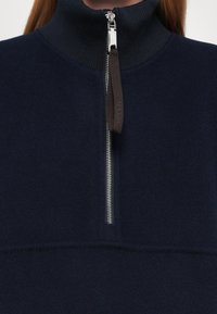 Primo piano di un indumento in pile blu navy con una zip argentata a metà e un pratico tirante in tessuto nero sul colletto a costine.