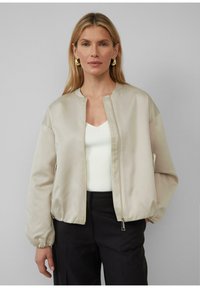 Beige satin bomberjack met een staande kraag, ritssluiting en elastische manchetten. Draagt over een witte top, gecombineerd met donkere broeken.
