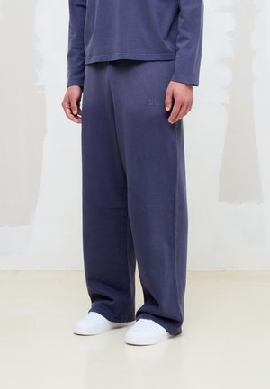 Persona vistiendo pantalones holgados azul marino y top de manga larga a juego con zapatillas blancas, de pie sobre un suelo llano frente a una pared clara.