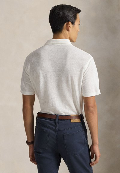 Άνδρας που φοράει λευκή πόλο μπλούζα με κοντά μανίκια, φορεμένη μέσα σε σκούρο μπλε παντελόνι με καφέ δερμάτινη ζώνη, εμφανιζόμενος από πίσω.