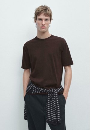 Młody mężczyzna z krótkimi włosami, ubrany w ciemnobrązową koszulkę i czarne spodnie, z paskiem swetra w paski związanego wokół talii, stoi na tle jasnej ściany.