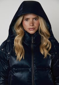 Giovane donna con lunghi capelli biondi che indossa un giubbotto imbottito blu scuro con cappuccio lucido, rivolta in avanti con un'espressione neutra.