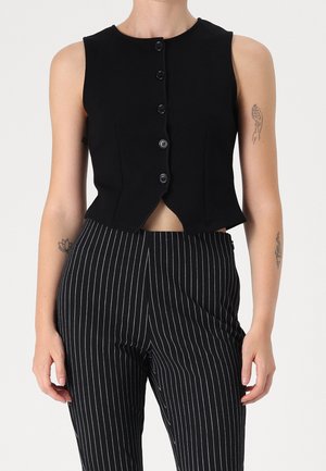 Gilet noir sans manches avec boutons, associé à un pantalon noir taille haute à fines rayures blanches verticales, porté par une personne tatouée.