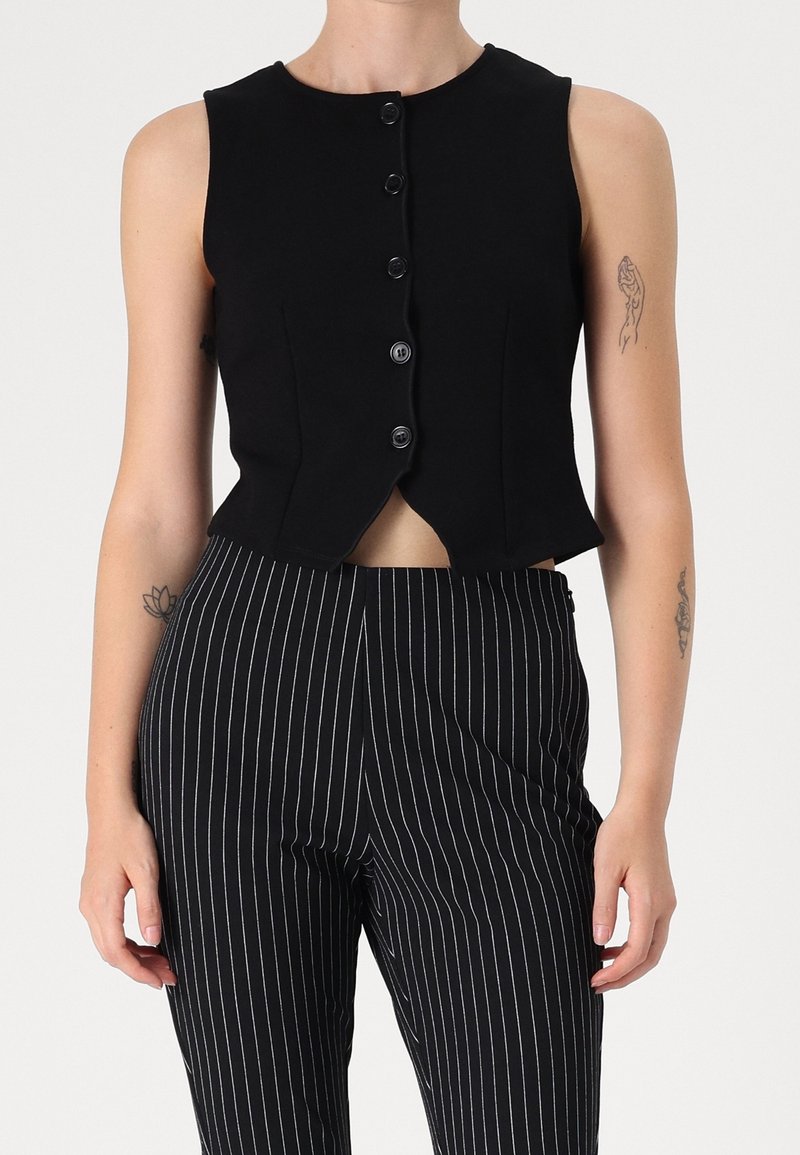 Gilet noir sans manches avec boutons, associé à un pantalon noir taille haute à fines rayures blanches verticales, porté par une personne tatouée.