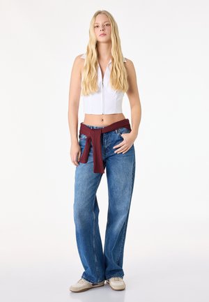 Blondynka stojąca z ręką w kieszeni, ubrana w bezrękawnik biały krótki top na zamek, luźne niebieskie jeansy, beżowe sneakersy oraz bordowy sweter zawiązany w talii.