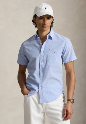 Joven con gorra blanca, camisa de manga corta azul claro a rayas con logo en el bolsillo, pantalones blancos y reloj de pulsera marrón, de pie con una mano en el bolsillo.