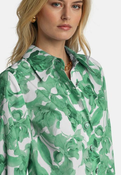 Blonde Frau, die ein weißes Hemd mit einem großen grünen Blumenmuster, goldene Creolen und eine goldene Kettenhalskette trägt.