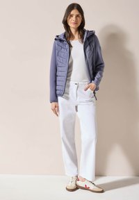 Gilet imbottito viola chiaro con cappuccio, abbinato a una camicia bianca e pantaloni bianchi a gamba larga. Sneakers beige con accenti rossi completano il look.