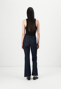 Γυναίκα με μακριά σκούρα μαλλιά στέκεται μπροστά σε έναν λευκό τοίχο, φορώντας μαύρο αμάνικο μπλουζάκι, σκούρα μπλε φαρδιά τζιν και μαύρα επίπεδα παπούτσια.