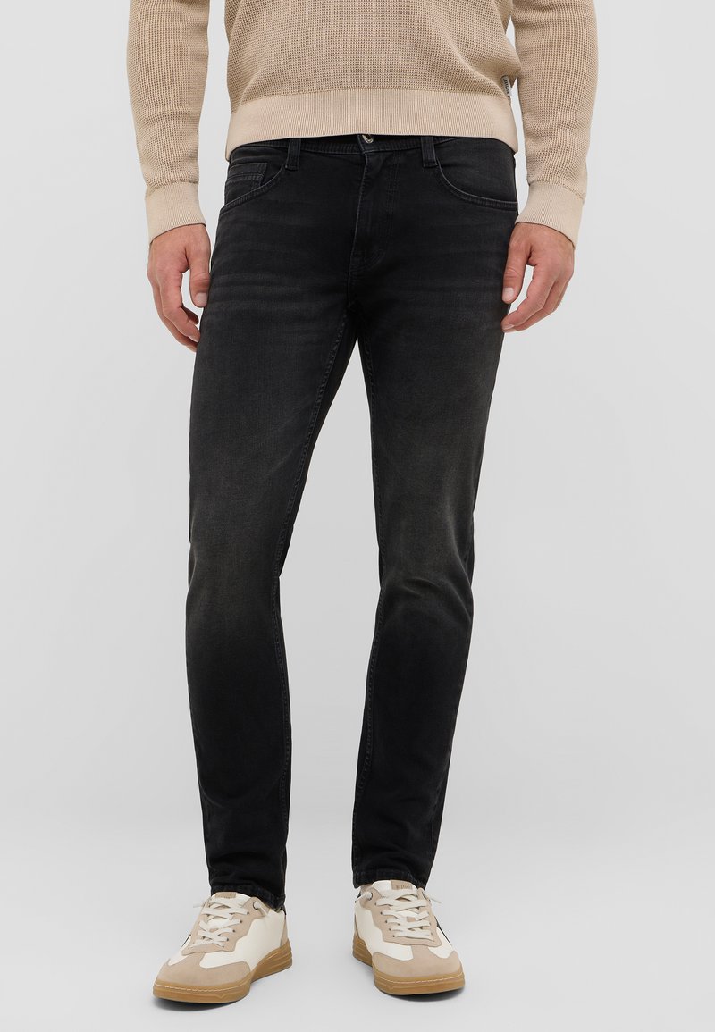 Fekete, slim fit farmernadrág, amely öt zsebbel rendelkezik és enyhén kopott textúrájú. Világos színű sportcipővel viselve.