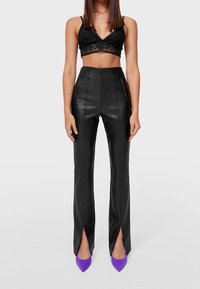 Zwarte faux leren high-waisted broek met voorlepen en contrasterende stiksels, gecombineerd met een zwart kanten crop top en paarse puntige hakken.