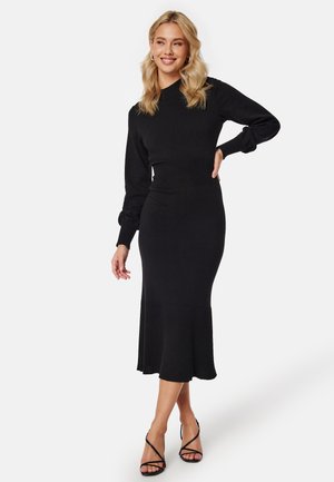 Robe en maille noire avec un corsage ajusté, des détails côtelés aux poignets et à l'ourlet, et une jupe évasée. Assortie à des sandales noires à lanières.