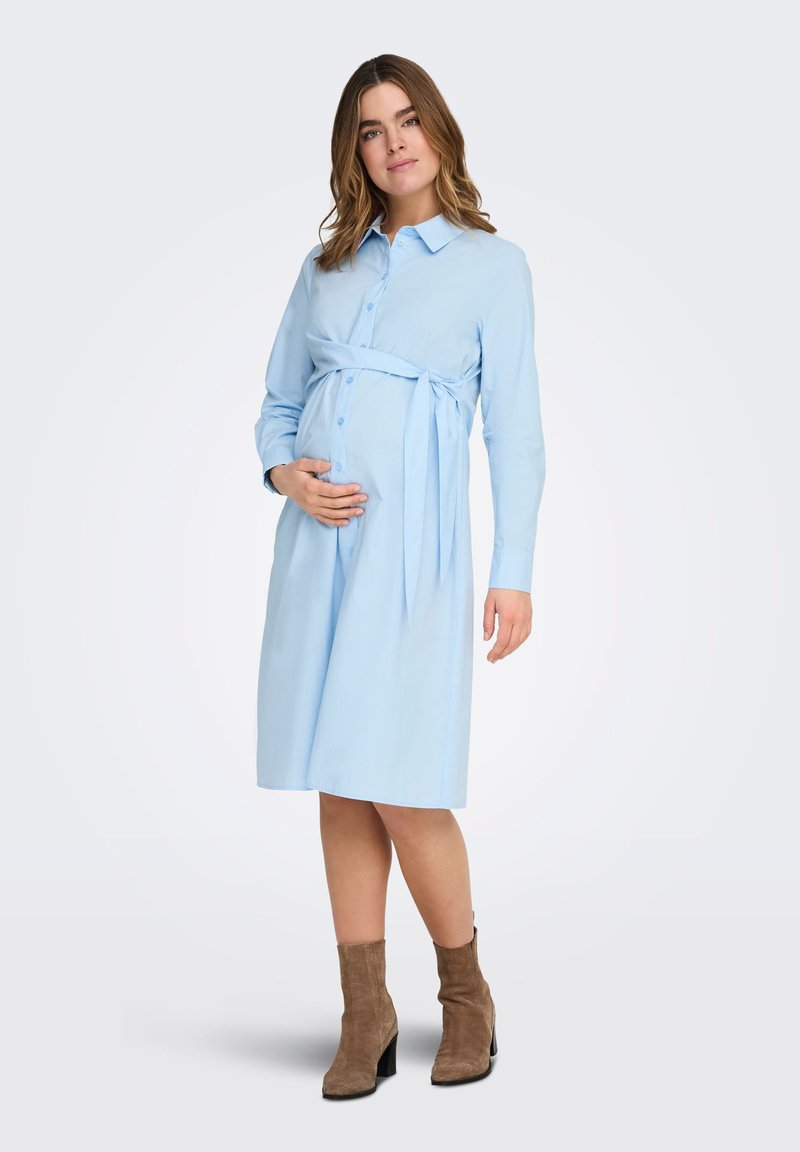 Έγκυος γυναίκα που στέκεται, φορώντας ένα ανοιχτό μπλε φόρεμα με κουμπιά και ζώνη στη μέση, μαζί με καφέ μποτάκια μέχρι τον αστράγαλο, κρατώντας την κοιλιά της.