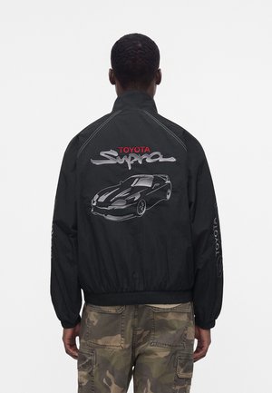 Mand med ryggen til iført sort jakke med broderet Toyota Supra-logo og bilgrafik, kombineret med camouflage cargobukser. Hvid baggrund.