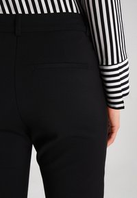 Des pantalons noirs ajustés présentent un design épuré, avec une texture lisse et une poche arrière discrète. Une chemise rayée est visible au niveau de la manche.