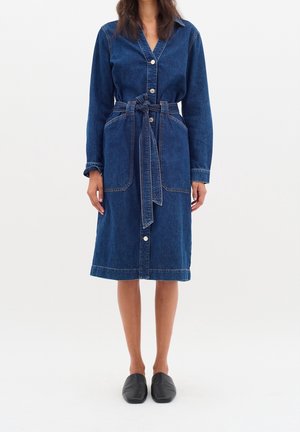 Kobieta ubrana w dżinsową sukienkę do kolan z długimi rękawami, zapinaną na guziki z przodu, z dużymi kieszeniami, paskiem do wiązania i czarnymi wsuwanymi butami.