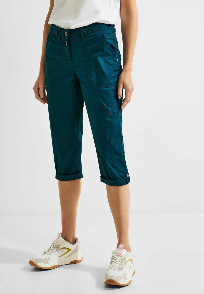 Personne portant un pantalon court bleu canard retroussé, des baskets blanches et une chemise blanche, debout sur un sol blanc uni.