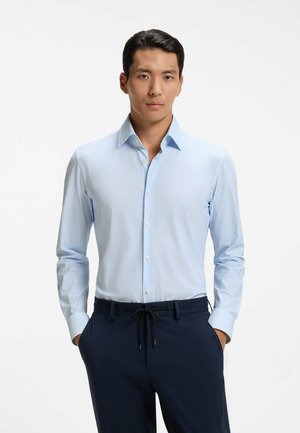 Mann trägt hellblaues Hemd mit Knöpfen und dunkelblaue Hose, steht mit den Händen in den Taschen vor weißem Hintergrund.