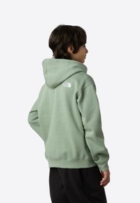 Zelena pamučna majica s kapuljačom, rebrastim manšetama i pojasom. Na gornjem dijelu leđa ima bijeli logo. Nošena uz crne hlače.