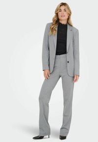 Femme avec de longs cheveux blonds portant un blazer gris et un tailleur-pantalon avec un top en dentelle noir et des talons aiguilles noirs, se tenant contre un fond blanc.