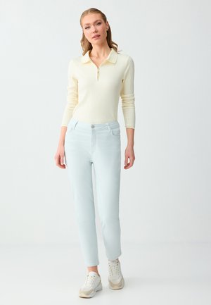 Kobieta w kremowej, prążkowanej bluzce z długim rękawem i zamkiem błyskawicznym, jasnoniebieskich dżinsach i białych trampkach, stoi na tle jednolitego jasnego tła.