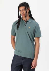 Zelená bavlnená polo tričko s krátkymi rukávmi, golierom a malým zlatým logom na ľavej hrudi. Nosené s tmavosivými džínsami.