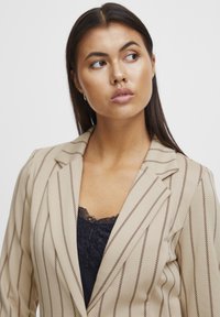 Beige Blazer mit Nadelstreifen, tailliertem Schnitt, strukturierten Revers und glatter Textur, übereinander getragen mit einem schwarzen Spitzen-Camisole.