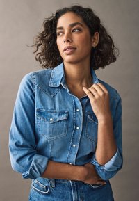 Jasnoniebieska koszula denimowa z zapięciem na guziki, dwoma kieszeniami na piersi i podwiniętymi rękawami. Oferuje klasyczny kołnierz i subtelne detale przeszyć.