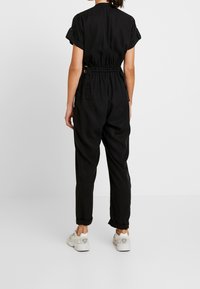 Zwart jumpsuit met korte mouwen, elastische taille en taps toelopende benen. Bevat opgerolde manchetten en zijzakken. Gemaakt met witte sneakers.