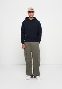Hanorac din bumbac bleumarin cu buzunar frontal tip kangaroo, asortat cu pantaloni cargo oliv, dotati cu multiple buzunare si mansete ajustabile. Tenisi albi.