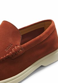 Brązowy zamszowy mokasyn wsuwany z białą gumową podeszwą i przeszyciami, z wytłoczonym logo "BOSS" na górnej części.