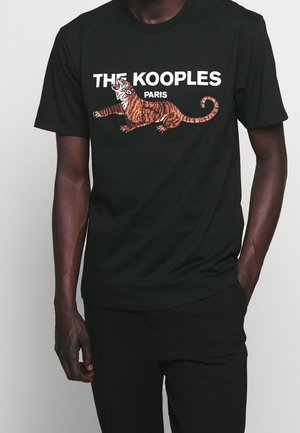 Mann in schwarzem T-Shirt mit einem hockenden orangefarbenen Tigermotiv und weißem Text "THE KOOPLES PARIS" auf der Brust.