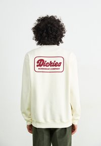 Cream-colored sweatshirt with a ribbed neckline and cuffs, featuring a maroon logo design that reads "Dickies WORKWEAR COMPANY" on the back.