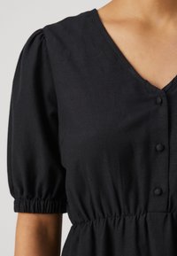 Μαύρο φόρεμα με V-λαιμόκοψη, κοντά φουσκωτά μανίκια και κουμπιά στο μπροστινό μέρος. Το ύφασμα φαίνεται λείο με μαζεμένη μέση.