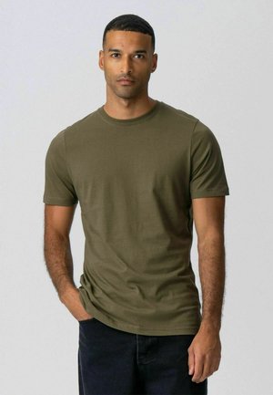 Lühikeste juustega mees, kes kannab oliivrohelise lühikese varrukaga t-särki ja tumedaid pükse, seisab ühe käega taskus, lihtsa tausta ees.