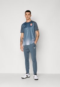 Sportbekleidungset bestehend aus einem kurzärmeligen, blauen T-Shirt mit Farbverlauf und Teamlogo sowie passenden, enganliegenden Jogginghosen. Mit gerippten Akzenten und Logodetails.