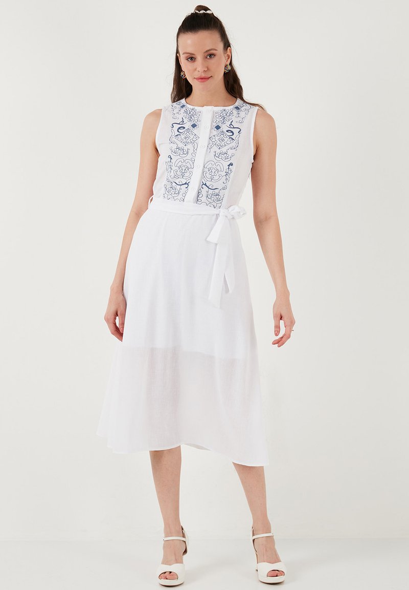 Weißes Leinenkleid mit ärmellosem Design, bestickten blauen Mustern auf der Vorderseite und einer Knoten-Taille. Der Rock ist halbtransparent und fließt bis zur Mitte der Wade.