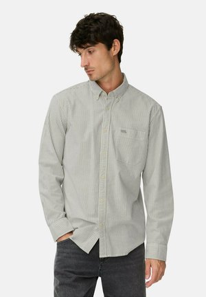 Young man wearing a light grey and white striped button-up shirt with long sleeves and dark jeans, hands in pockets, looking down left.