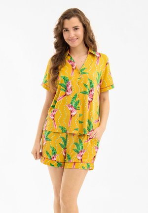 Mujer con camisa amarilla de manga corta abotonada y pantalones cortos a juego con estampado de pájaro rosa y hojas verdes, sonriendo con la mano en el bolsillo.