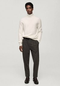 Pulover cu guler înalt de culoare crem, cu un model texturat, asortat cu pantaloni ajustați de culoare gri închis și cizme maro până la gleznă. Design simplu, modern.