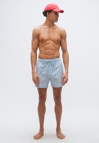 Μυώδης άνδρας χωρίς μπλούζα, φορώντας γαλάζια ριγέ σορτσάκια και κόκκινο καπέλο, όρθιος ξυπόλητος και στραμμένος προς τα αριστερά σε λευκό απλό φόντο.