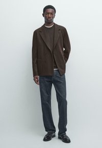 Hnědý vlněný dvouřadý blejzr přes tmavý svetr, doplněný tmavými džíny s rovným střihem a černými mokasínami. Minimalistický design, šité na míru.