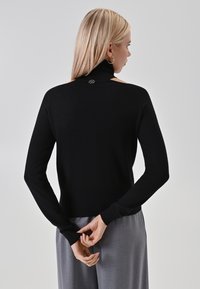 Maglione nero a maniche lunghe con collo alto e dettagli sulle spalle scoperte. Texture a maglia con un accento del logo sul retro vicino al colletto.