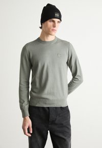 Groene gebreide trui met een ronde hals en geribbelde manchetten. Bevat een klein vierkant logo op de borst. Gecombineerd met een zwarte jeans en een zwarte beanie.