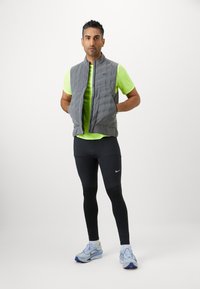 Grå vadderad väst över en ljusgrön sporttröja, kombinerad med svarta leggings och ljusblå sportskor med lila accenter.