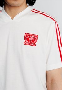 T-shirt branca da Adidas com riscas vermelhas nas mangas e um logótipo bordado em vermelho no peito. Tecido suave com um design desportivo e decote em V.