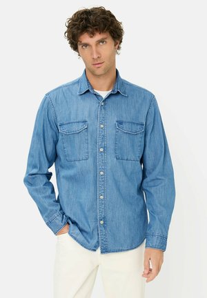 Mężczyzna o kręconych włosach, ubrany w niebieską koszulę z denimu zapiętą na guziki, na białej koszulce pod spodem i jasnych spodniach, stojący z jedną ręką w kieszeni.