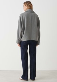 Giacca oversized in misto lana grigia con ampio colletto, vestibilità rilassata e dettaglio sulla cucitura posteriore, abbinata a pantaloni flare in denim scuro.