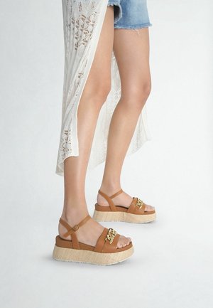 Sandalias de plataforma marrones con suela de corcho tejida, con tiras en el tobillo y un detalle de hebilla dorada. Usadas con pantalones cortos de mezclilla y un chal ligero.